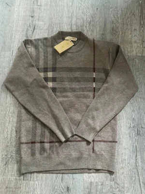 Burberry tröja  - Snygg beige tröja från Burberry med klassiskt rutmönster i grått och vinrött på framsidan. Rund halsringning och långa ärmar med ribbade muddar. Mjuk och tunn stickad kvalitet som känns lyxig och stilren. Perfekt för dig som gillar designerplagg. Slim fit 