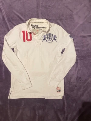 Vit långärmad rugbyskjorta Superdry - Säljer en vit långärmad rugbyskjorta från Superdry med tryckta detaljer i rött och blått, inklusive nummer 10 och ett lejonemblem på bröstet. Skjortan har klassisk krage och knappar vid halsen. Perfekt för dig som gillar sportig stil.