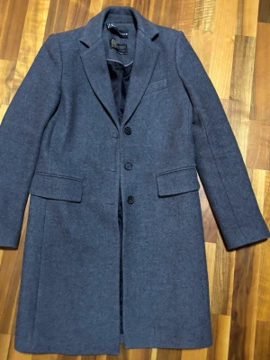 Klassisk grå kappa från Zara - Klassisk varm kappa i Manteco-ullblandning från ZARA  Melerad mörkgrå färg, strl XS, men den kan också passa en S beroende på hur man vill den ska sitta. Knäppning med knappar, fickor med flärpar. 65% Manteco ull, gjord i Marocko.   Nypris ca 1 500kr.