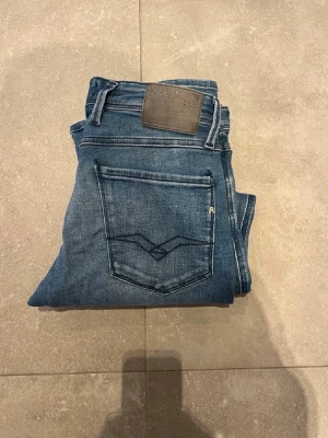 Replay blå jeans med klassisk passform - Säljer ett par snygga blå jeans från Replay med klassisk femficksdesign och tydliga sömmar. Jeansen har en normal passform och är tillverkade i mjukt jeansmaterial med Replay-logga på bakfickan och läderpatch i midjan. Perfekta för dig som gillar stilrena och tidlösa jeans.