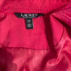 Ralph lauren trenchcoat med dubbelknäppning - Snygg ceriseröd trenchcoat från Ralph Lauren med klassisk dubbelknäppning och axelklaffar. Jackan har fin ceriseröd färg, långa ärmar och är tillverkad i ett slätt material. Välarbetat foder med svagt mönster. Perfekt för dig som vill sticka ut med färg i vår eller höst. Något smutsig i ärmslut men aldrig tvättad o använt fåtal gånger. Fläck på skärp men syns inte på. Passar bäst en 40-44. Jag är 169 o liten M så för stor tyvärr.