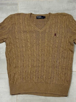 Beige stickad tröja från Polo Ralph Lauren - Säljer en beige stickad tröja från Polo Ralph Lauren i storlek L. Tröjan har klassiskt kabelstickat mönster, v-ringad hals och långärmade ribbade muddar. Ikoniska Polo-loggan broderad på bröstet i rött. Perfekt för dig som gillar stilrena och tidlösa plagg.