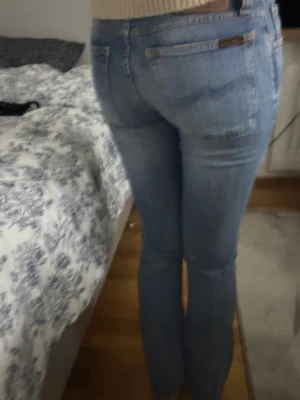 Ljusblå jeans från Nudie Jeans - Säljer ett par ljusblå jeans från Nudie Jeans med klassisk femficksdesign och snygga kontrastsömmar. Jeansen har en rak passform och är tillverkade i mjukt bomullsmaterial. Märkespatch i skinn baktill och diskret logga vid bakfickan.