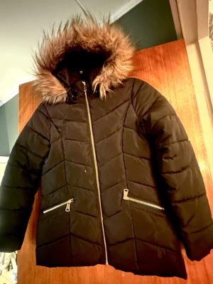 Svart pufferjacka med fuskpäls - Svart pufferjacka med fluffig fuskpäls på huvan.  Perfekt för kalla dagar då den är varm och skön. Jackan är i mycket fint skick då dottern inte använt den många gånger, hittar inga fel när jag kollat. Stl 158