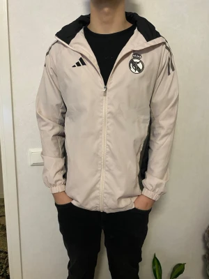 Beige Real Madrid vindjacka  - Säljer en beige vindjacka från Adidas X Real Madrid. Jackan har svarta detaljer på axlarna och dragkedja framtill. Perfekt för dig som gillar sportig stil och vill visa ditt lag. Den har använts några gånger och har inga defekter. Den är storlek M,  ställ frågor vid minsta fundering 😊✨ Ny pris: 1152kr
