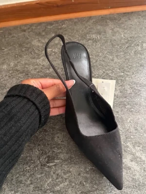 Svarta pumps med spetsig tå från H&M - Stilrena svarta pumps från H&M med spetsig tå och smal klack. Skorna har en öppen häl med rem och är tillverkade i syntetmaterial med mockakänsla. Perfekta för dig som vill ha en clean och trendig look.
