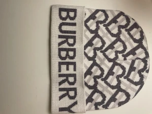 Vit och grå Burberry mössa - Säljer en snygg mössa från Burberry i vit och grå färg med stort BURBERRY-tryck på uppviket och mönster av B-bokstäver över hela mössan. Stickad i mjukt material som håller dig varm och stilren under kalla dagar.