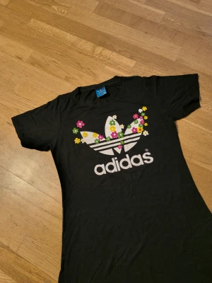 Svart Adidas klänning med blommor - Svart långklänning från Adidas med korta ärmar och stort vitt Adidas-tryck framtill dekorerat med färgglada blommor i rosa, gul och grön. Klänningen har en rak och enkel form, tillverkad i mjuk bomull som känns skön mot huden.