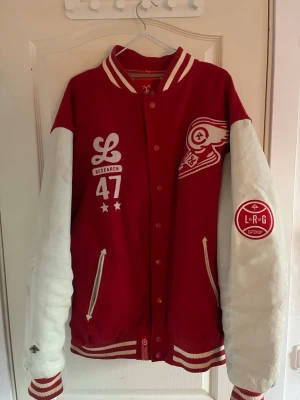  LRG Varsity jacka - Detroit red wings - Säljer en fet vintage röd och vit varsityjacka från LRG i storlek XXL. Jackan har broderade patches, vita ärmar, ribbade muddar och krage med ränder, samt quiltat beige foder. Materialet är 90% ull och 10% polyester med polyesterfoder. Snygga detaljer med tryck och knappar framtill. Jätte y2k streetstil på den. Men passar även Detroit red wings nhl fansen.