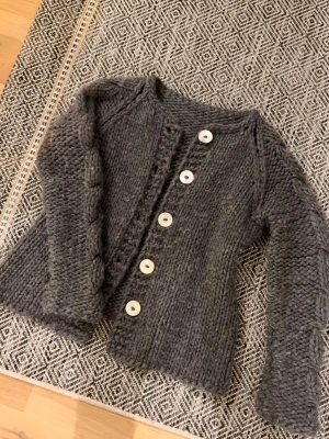 Mörkgrå stickad kofta med pärlemorknappar - Säljer en chunky mörkgrå stickad kofta med stora pärlemorknappar framtill. Koftan har långärmade ärmar med flätad detalj och rund hals. Perfekt för lager-på-lager och att mysa i under kyliga dagar.