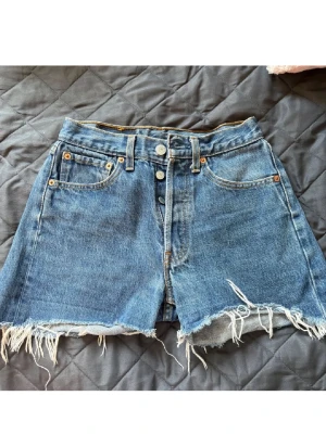 Levis Blå denimshorts med fransig kant - Säljer ett par blå Levis denimshorts med hög midja och fransig, rå kant längst ner. Klassisk femficksmodell med knappar framtill och slitna detaljer för en cool vibe. Perfekta för varma dagar och chill häng.