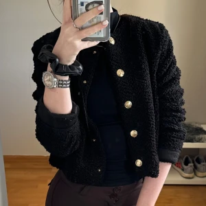 Svart teddyjacka med guldknappar - Säljer en svart teddyjacka med snygga guldknappar framtill. Jackan har en croppad passform och långärmade ärmar. Perfekt för dig som vill ha en cool och trendig look med extra textur och detaljer.