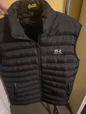 Svart väst från Under Armour - Säljer en svart dunväst från Under Armour med quiltad design och dragkedja framtill. Västen har två praktiska fickor med dragkedja och en hög krage som ger extra värme. Perfekt för lager-på-lager och sportiga outfits. Storlek M, knappt använd!