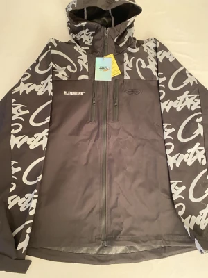 Corteiz ELITEWORK windbreaker - Svart vindjacka från ELITEWORK med grått grafiskt mönster på ärmar och huva. Jackan har två vertikala dragkedjor framtill, huva och tryckt logga på bröstet. Materialet är syntet och jackan har en loose passform. Storlek: L/XL. Pris kan diskuteras