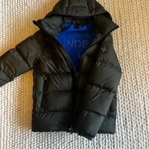 Grön pufferjacka från J.Lindeberg - Snygg mörkgrön pufferjacka från J.Lindeberg med blått foder, huva och två dragkedjeförsedda fickor. Jackan har ett stilrent emblem på ärmen och quiltad design som ger en cool look. Perfekt för kalla dagar och enkel att matcha med dina favoritkläder.