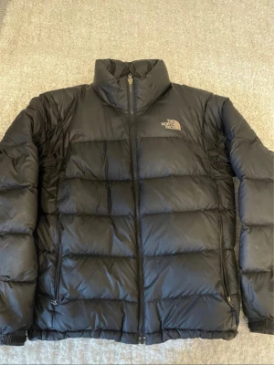 Svart pufferjacka från The North Face - Säljer en svart pufferjacka från The North Face med hög krage och dragkedja framtill. Jackan har två sidofickor med dragkedja och ett stilrent, quiltat mönster. .