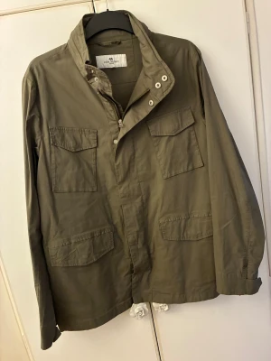 Grön poly jacka från John Henric - Snygg olivgrön poly jacket från John Henric med fyra stora fickor framtill och hög krage. Jackan har dragkedja och knappar. Använd 1 gång och skicket är helt nytt. Pris kan diskuteras!