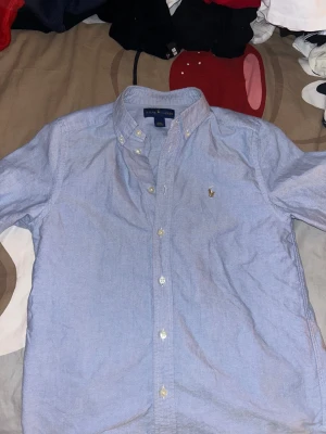 Blå skjorta från Ralph Lauren - Snygg ljusblå skjorta från Ralph Lauren med klassisk button-down krage och broderad logga på bröstet. Skjortan har långa ärmar och vita knappar framtill. Perfekt för dig som gillar stilrena och tidlösa plagg.