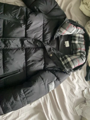 Svart pufferjacka från Burberry - Svart pufferjacka från Burberry med avtagbara ärmar, vilket gör den till en väst också. Jackan har ett stort Burberry London England-märke på ärmen och ett rutigt foder i huvan. Materialet är polyester och dun, perfekt för kalla dagar.