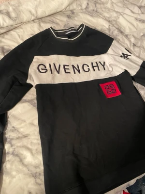 Svart och vit Givenchy sweatshirt - Säljer en svart och vit sweatshirt från Givenchy med broderad logga på bröstet och texten 'PARIS' på ärmen. Tröjan har en röd patch med Givenchy-symbolen och ribbad krage med vit rand. Perfekt för dig som gillar exklusiva streetwear-vibes.