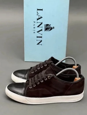 Lanvin sneakers i mocka och skinn - Snygga sneakers från Lanvin i mörkbrun mocka med svart skinn på tån och vit platt sula. Skorna har rund tå, snörning och diskret design som passar till många stilar. Perfekta för dig som gillar exklusiva och stilrena sneakers.