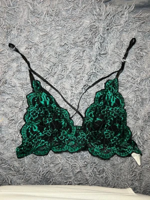 Grön spets bralette med justerbara band - Säljer en snygg bralette med grön och svart spets, blommigt mönster och tunna justerbara axelband. Toppen är ärmlös och har en triangelformad kupa med dekorativ spetskant. Perfekt att styla under en skjorta eller som statement-piece. Står XXL men skulle mer säga XL