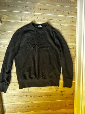 Svart sweatshirt från Moncler - Svart sweatshirt från Moncler med broderad logga på bröstet och en ton-i-ton patch på ärmen. Tröjan har rund hals, ribbade muddar och är långärmad. Materialet är mjuk bomull som känns skönt mot huden. Perfekt för dig som gillar stilrena och exklusiva plagg. Skick 8/10. Nypris: ca 6000kr