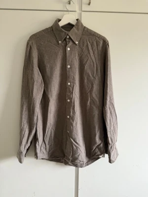 Brun skjorta från Morris Heritage - Snygg brun skjorta från Morris Heritage med klassisk krage och vita knappar. Skjortan är långärmad och tillverkad i ett mjukt bomullsmaterial som ger en chill och stilren look. Perfekt för dig som gillar enkel design och vill ha något som funkar året runt.