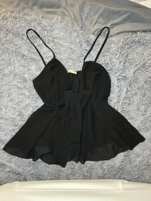 Svart linnetopp med rosett från New Style - Säljer en svart linnetopp från New Style Italy med tunna axelband och själkyten rosett framtill. Toppen har en flowy och luftig passform med omlottliknande lager och är tillverkad i ett lätt syntetmaterial. Perfekt för dig som gillar stilrena och feminina detaljer. Tyget är väldigt genomskinligt, vid brösten är det mer tyg. Men fortfarande lite egenomskinligt 