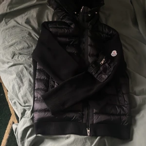 Moncler cardigan  - Svart pufferjacka från Moncler med huva och ribbade stickade ärmar. Jackan har dragkedja framtill och en ficka med dragkedja på ärmen där Moncler-loggan sitter. Materialet är en mix av polyester och stickat, perfekt för kyliga dagar.
