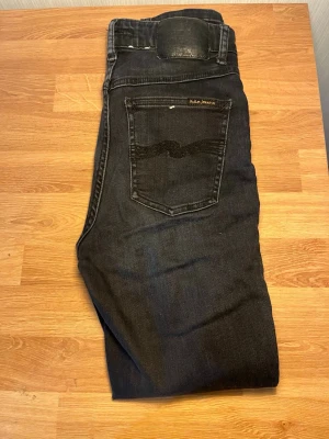 Svarta jeans från Nudie Jeans - Säljer ett par svarta jeans från Nudie Jeans med klassisk femficksdesign och snygg broderad bakficka. Jeansen har en mörk tvätt, silverfärgade nitar och dragkedja i gylfen. Märkespatch i svart skinn baktill. Perfekt för dig som gillar stilrena och tidlösa jeans. W28 l26