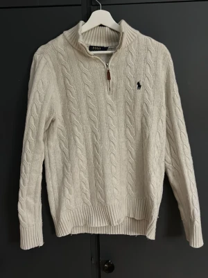 Vit stickad tröja Polo Ralph Lauren - Snygg vit stickad tröja från Polo Ralph Lauren med klassiskt kabelmönster och broderad logga på bröstet. Tröjan har en ribbad krage och half zip-dragkedja framtill. Perfekt för dig som gillar stilrena och tidlösa plagg.