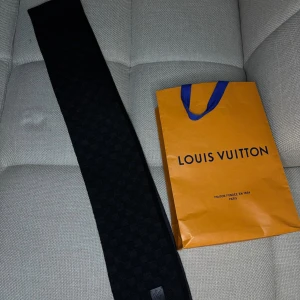 Svart Louis Vuitton halsduk - Säljer en svart halsduk från Louis Vuitton med diskret rutigt mönster och en liten logotyp-patch. Halsduken har en stilren och tidlös design som passar perfekt till jacka eller kappa. Kommer med originalpåse från Louis Vuitton.