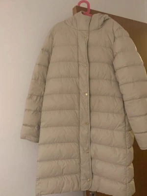 Beige lång pufferjacka Part Two - Säljer en beige pufferjacka från Part Two i storlek 36. Jackan är lång, har huva och guldfärgade knappar framtill. Perfekt för kalla dagar och har en clean, stilren look som är lätt att matcha med olika outfits.