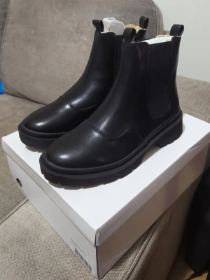 Nelly stövlar svarta  - Snygga svarta chelsea boots från Nelly med grov sula och rund tå. Utsidan är i slätt skinn och insidan är fodrad med mjukt beige pälsmaterial för extra värme. Bootsens elastiska paneler gör dem enkla att ta på och av. Perfekta för höst och vinter! Storlek 38