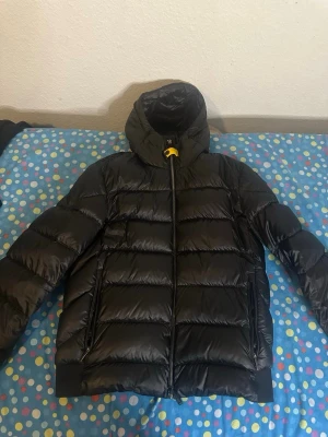 Svart dunjacka Parajumpers med huva - Säljer en svart dunjacka från Parajumpers med glansig finish och huva. Köpte jackan förra vintern och har nu köpt en ny jacka så kommer inte behöva denna något mer. Jackan är fräsch och har inga defekter och är äkta Köpt från NK