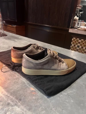 Lanvin sneakers i brun mocka - Säljer ett par Lanvin sneakers i brun mocka med snörning och rund tå. Skorna har en beige gummisula och en blank, bronsfärgad tå i syntetmaterial som ger en unik look. Perfekta för dig som gillar stilrena och exklusiva sneakers.