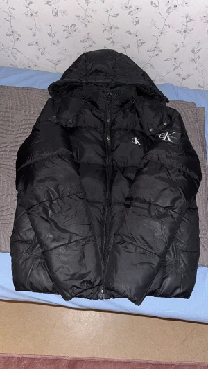 Svart pufferjacka från Calvin Klein Jeans - Svart pufferjacka från Calvin Klein Jeans med huva och dragkedja framtill. Jackan har broderad CK-logga på bröstet och är tillverkad av 100% återvunnen polyester. Vattenavvisande och fodrad för extra värme, perfekt för kyliga dagar.