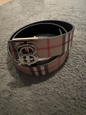 Rutig beige bälte med metallspänne - Säljer ett snyggt bälte från Burberry med klassiskt rutig design i beige, rött och brunt. Bältet har ett stort metallspänne med Burberrys ikoniska B-logga. Perfekt för att lyfta din outfit med lite lyxig vibe.