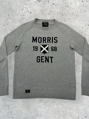 Grå stickad tröja Morris Gents M - Snygg grå stickad tröja från Morris Gents i storlek medium. Tröjan har svart broderad text och ett emblem på bröstet, samt ribbade muddar vid ärmslut och nederkant. Perfekt för dig som gillar klassisk stil med en modern touch. Hör av er för minsta fundering,💨