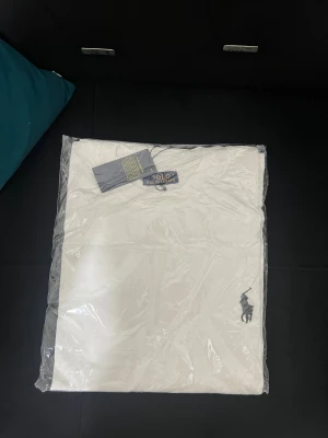 Vit t-shirt från Polo Ralph Lauren - Vit t-shirt från Polo Ralph Lauren med klassisk rund hals och korta ärmar. T-shirten har den ikoniska Polo-loggan broderad i mörk färg på bröstet. Tillverkad i mjuk bomull för skön komfort och stilren look.