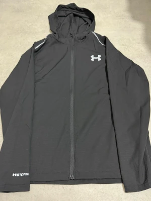Svart vindjacka Under Armour M - Svart vindjacka från Under Armour med huva och hel dragkedja. Jackan har vita detaljer på axlarna och logga på bröstet. Materialet är lätt och syntetiskt, perfekt för träning utomhus. Modellen är långärmad och har HeatGear-teknologi för bra ventilation.
