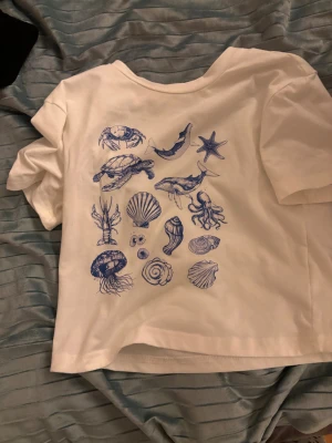 Vit t-shirt med blått havsmotiv - Vit t-shirt med coola blå tryck av havsdjur och snäckor på ryggen. Motiven inkluderar sköldpadda, val, haj, krabba, sjöstjärna, manet och olika snäckor. T-shirten har korta ärmar och en avslappnad passform. Perfekt för dig som gillar havet och unika prints. ( den är cropped så man kan ha på sig den vid 152 också )