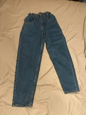 Blå högmidjade jeans med rynkad midja - Säljer ett par blå jeans med rynkad, hög midja och två knappar framtill. Jeansen har klassiska fickor och gul kontrastsöm. Modellen är rak och något loose, med dragkedja och bälteshällor. Perfekt för dig som gillar retrovibe och bekväm passform. Byxorna har även en kort längd 