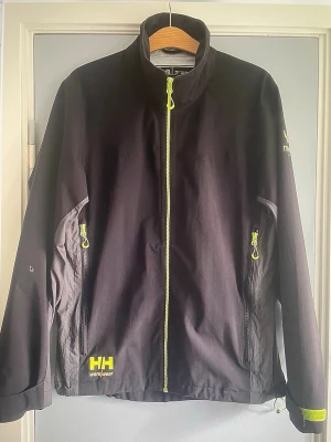 Svart vindjacka Helly Hansen Workwear - Svart vindjacka från Helly Hansen Workwear med neongula dragkedjor och detaljer. Jackan har hög krage, två sidofickor med dragkedja och justerbara ärmslut. Materialet är slitstark polyester och jackan har en sportig vibe med tydlig logga framtill.