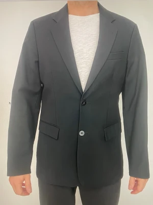 J.Lindeberg kavaj - Säljer en mycket fin och stilren kavaj från J.Lindeberg Stockholm i nyskick. Klassisk modell med två knappar, perfekt både för jobb, fest och mer uppklädda tillfällen. Tidlös design i hög kvalitet. Detaljer: * Märke: J.Lindeberg  * Storlek: 48  * Material: 100% ull  * Färg: svart / mycket mörk antracit  * Knäppning: 2 knappar  * Passform: modern och välsittande  Mått (ca): * Bröstvidd: 53 x 2 cm  * Midjevidd: 48 x 2 cm  * Stussvidd: 55 x 2 cm  * Axel till axel: 45 cm  * Ärmlängd från axel: 65 cm