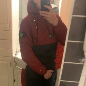 Röd och svart vindjacka Stone Island - Säljer en snygg tvåfärgad vindjacka från Stone Island med huva och snörning. Jackan är röd upptill och svart nedtill, har lång ärm och klassisk patch på vänster arm. Perfekt för dig som vill ha streetstyle med ikonisk look.