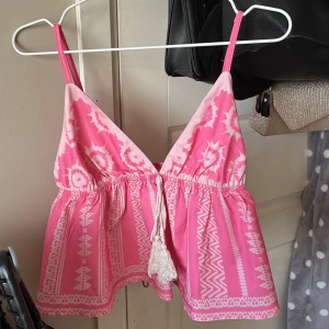 Rosa mönstrad linnetopp - Supersöt rosa linnetopp med vitt etnomönster och smala axelband. Toppen har v-ringning, knytning med tofsar framtill och en luftig, flowy passform. Perfekt för varma dagar och ger en riktigt somrig vibe 💕🤍