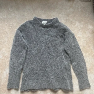 Grå fluffig stickad tröja H&M - Supermjuk och fluffig grå stickad tröja från H&M i ull- och alpackablandning. Tröjan har en rak passform, lång ärm och lite högre hals. Perfekt för kyliga dagar när du vill vara både varm och snygg.deras exklusiva kollektion 