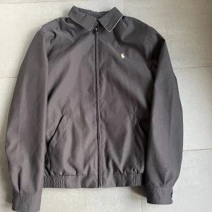 Svart jacka från Polo Ralph Lauren - Nytt pris är ungefär 2699 Svart jacka från Polo Ralph Lauren med klassisk krage, dragkedja framtill och diskret broderad logga på bröstet. Jackan har två snedställda fickor och elastisk midja. Fodret är svart med rutigt mönster. Knappt använd har du frågor så är det bara att ställa